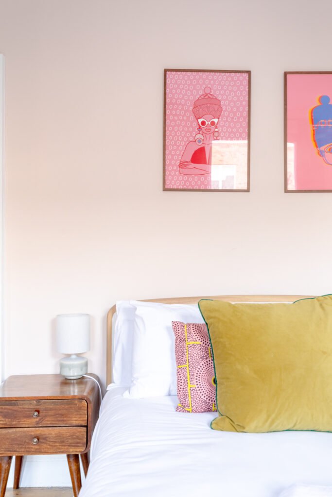 Partial view of a bed, a bedside table in a pink coloured bedroom, with pink images on the wall.