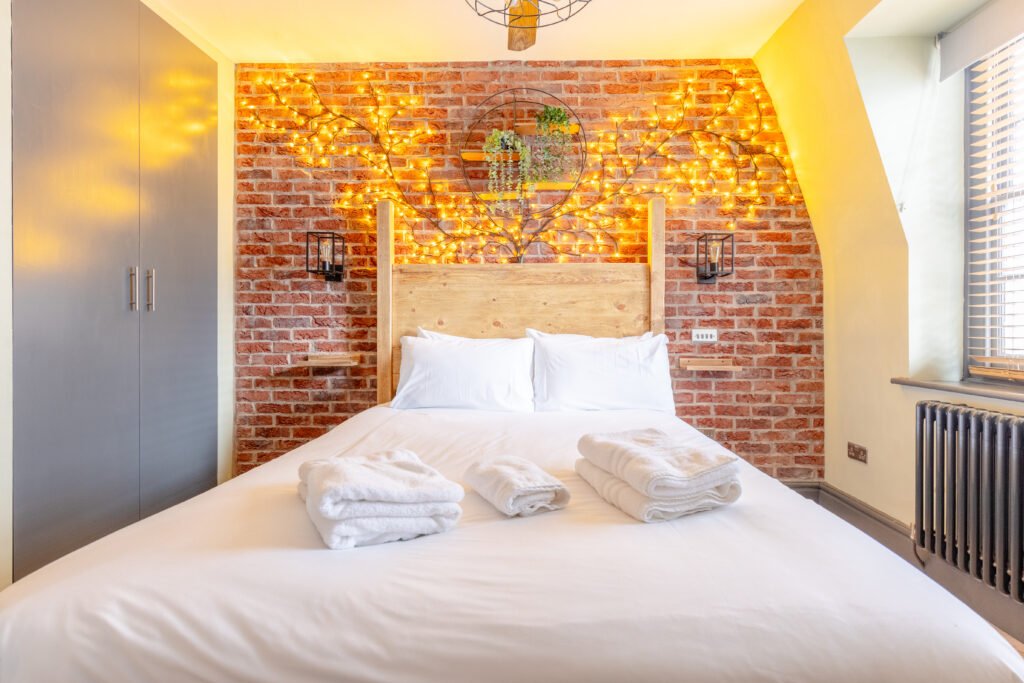 A bedroom photo for property photography, showing a bed from front, white sheets and behind is a brick wall with warm yellow coloured lighting.