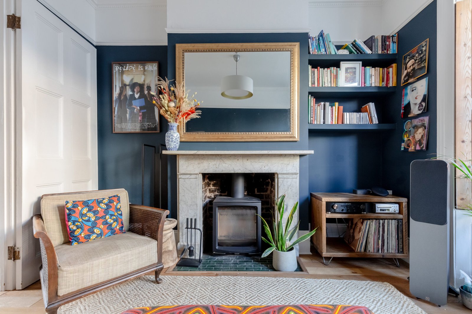 A cosy living room with a small wood stove in the fireplace, an armchair, dark blue walls and bookshelves on the wall.